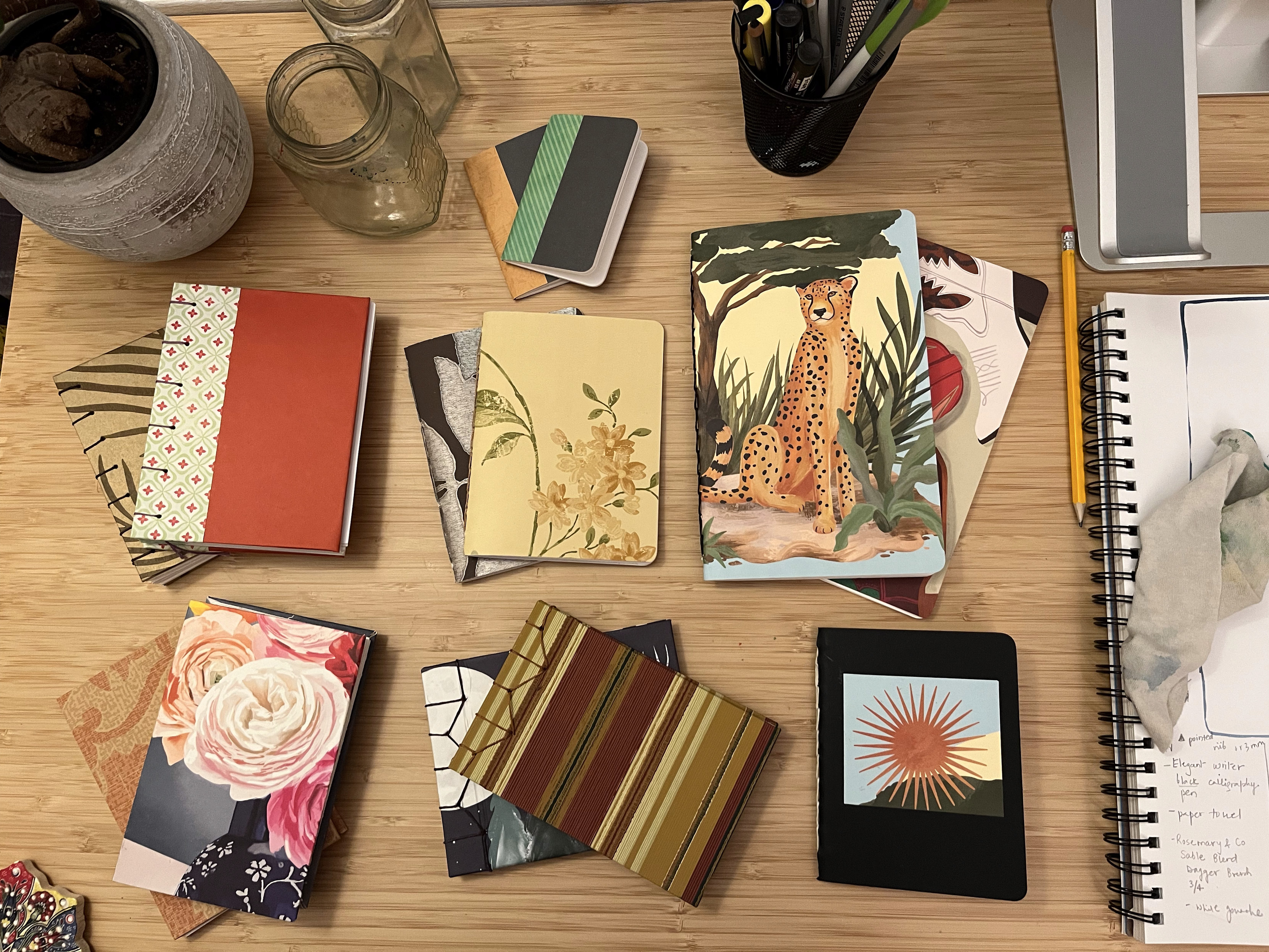 A collection of various handmade notebooks on a bamboo desk. In the top row are two small pamphlet stitch books. In the middle row are two Coptic stitch books, two medium sized pamphlet stitch books, and two large pamphlet stich books. The bottom row has two accordion style books, two stab bind books, and one smaller pamphlet stitch books.