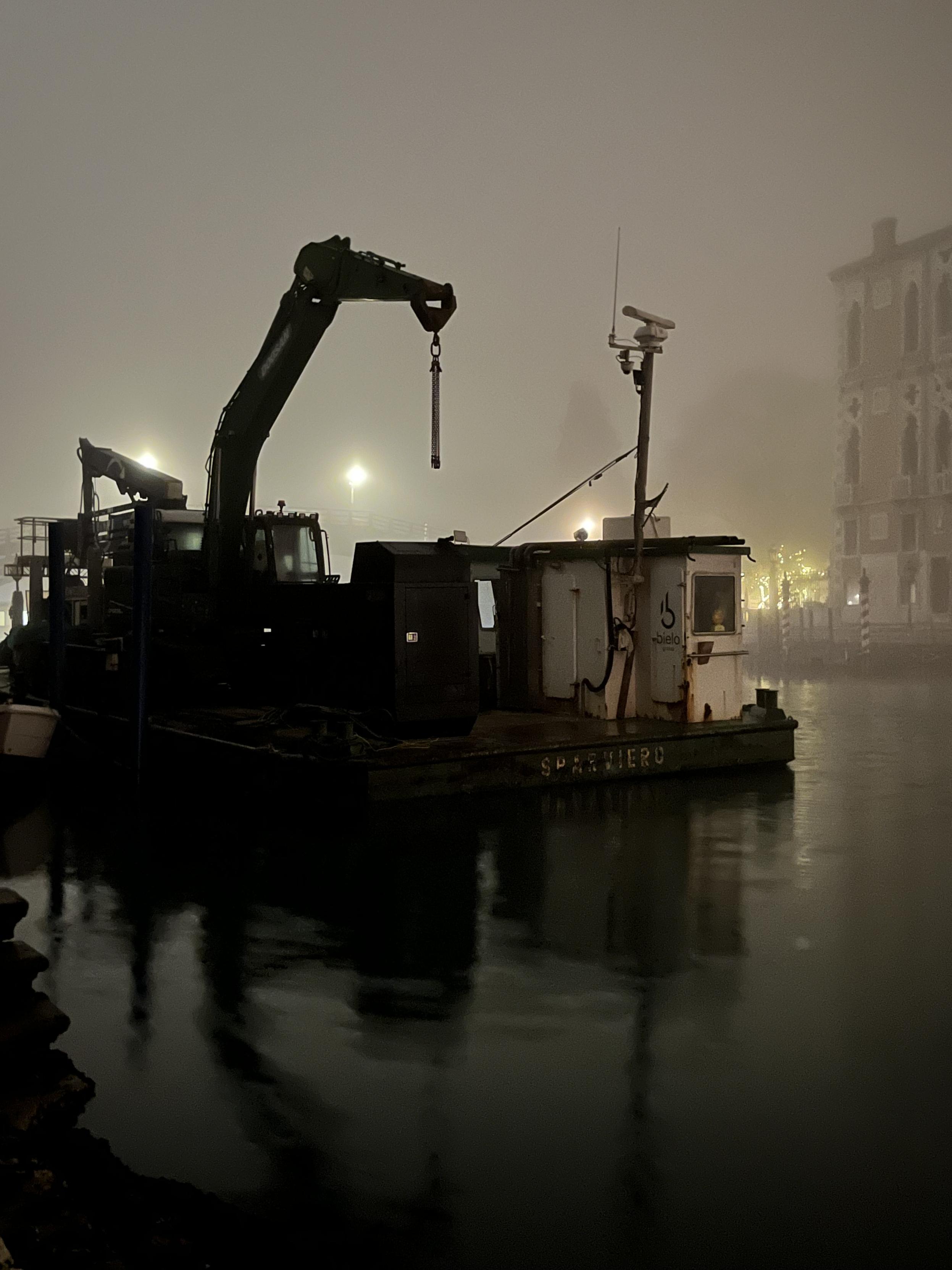 Dark and moody colour photo, taken on a foggy night, of a big platform with building equipment on it floating on the Canal Grande. The most immediately visible objects are a dingy white metal hut at the canal edge of the platform, and a big black crane with a chain hanging from the end of its tall arm, and there are other chunky black structures in deep darkness rendering them indistinct. The scene is lit from behind by three round streetlights which glow through the dense grey fog, and a mess of fairy lights in a garden on the opposite side of the canal. The water in the canal is black and very gently rippled, and the platform and its cargo leave even deeper black reflections. The feeling in the photo is mysterious and a little bit spooky. 