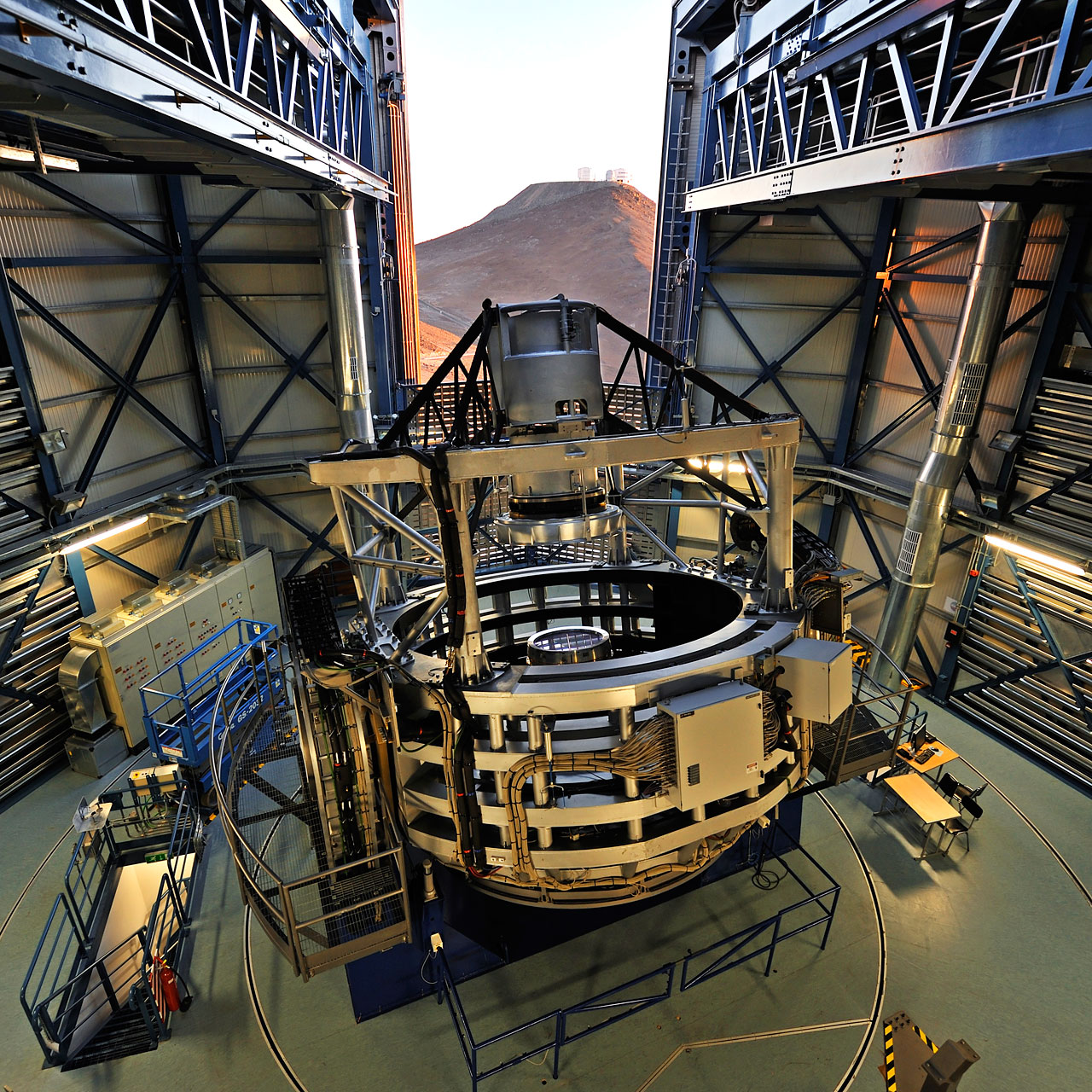 An image of a telescope inside its dome. The telescope is an open cylindrical structure with a 4-m wide primary mirror. The dome is partially open; it's daytime outside, and the dome's opening reveals a few other telescopes perched on a distant hill.