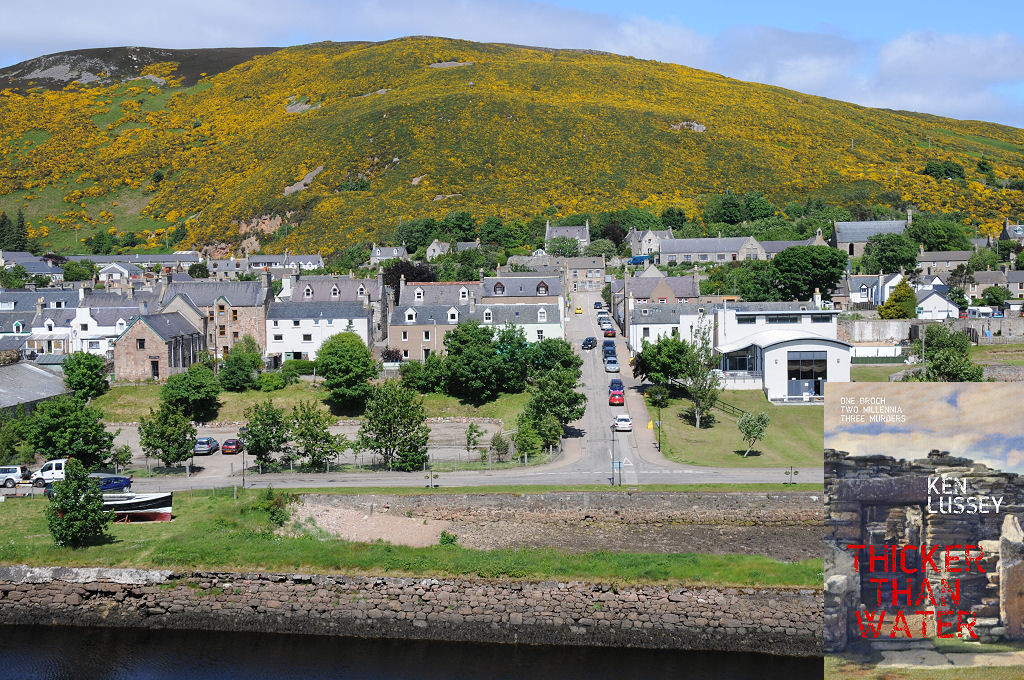 The image shows a view down and across a river which runs along the bottom of the frame. Above the stone riverbank and an open area with trees and a road is a settlement of mainly stone two and three storey buildings extending in a band across the frame. Beyond it, a hillside covered in yellow gorse rises to the skyline. The scene is in sunlight. The front cover of ‘Thicker Than Water’ is shown in the bottom right corner.