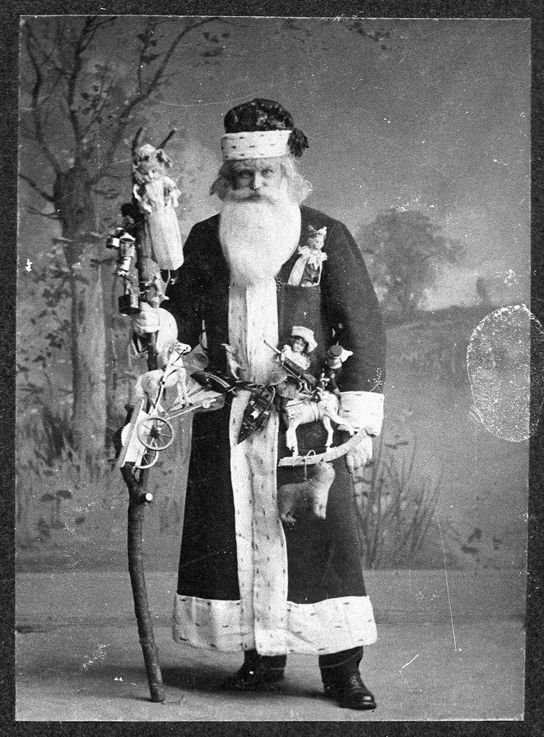 A black and white photo of a man dressed in a long coat with white trimmings, a santa-hat, and a big white beard. He has a wooden staff with him, which multiple toys are attached to. He is posed in front of a countryside-scene backdrop, with a tree to the left and field to the right.
Photo caption on NMS reads: 'Mr Henry Watson, dressed up as Father Christmas in Burntisland, Fife, in 1909. Reference SLA 63.16.11A. Credit: Donated by Miss C. Watson, photographer unknown.'