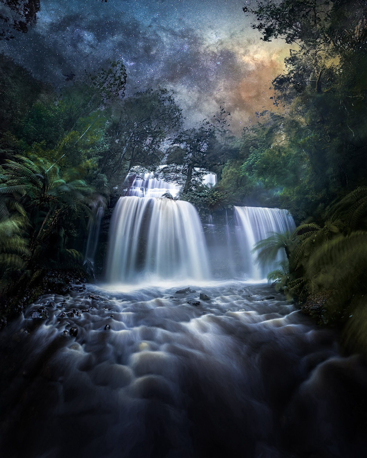 The scene is focused on the full flow of Russell Falls, rimmed by prehistoric ferns and subalpine rainforest. The foreground has intense flow running over rocks, with significant spray from the falling water. In the canopy, the galactic core of the Milky Way sets behind the zenith of the falls.