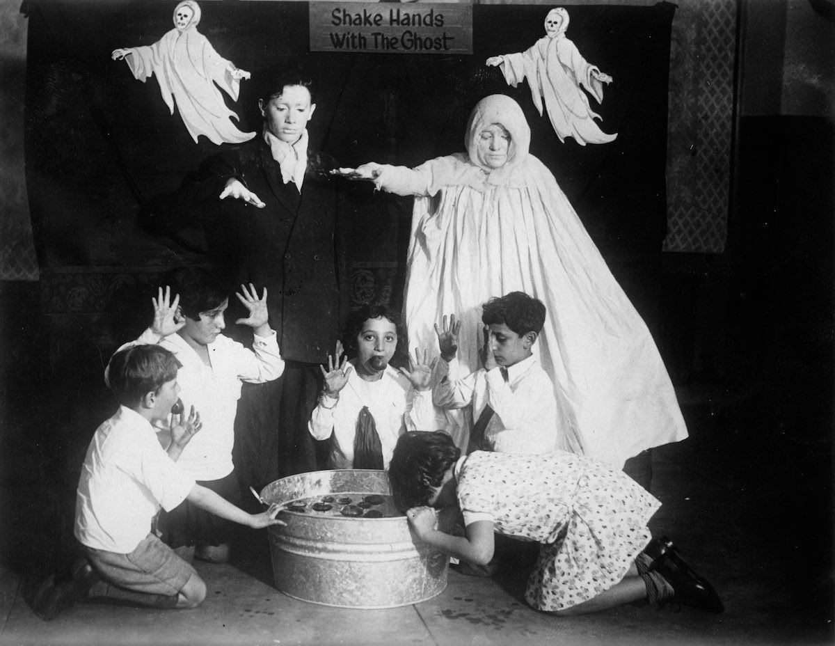 A black-and-white photo showing a group of kneeling children apple bobbing around a water-filled tub. Two adults dressed as ghosts are behind them before a screen decorated with two flying, skull-headed ghosts and a notice saying 'Shake hands with the ghost'.