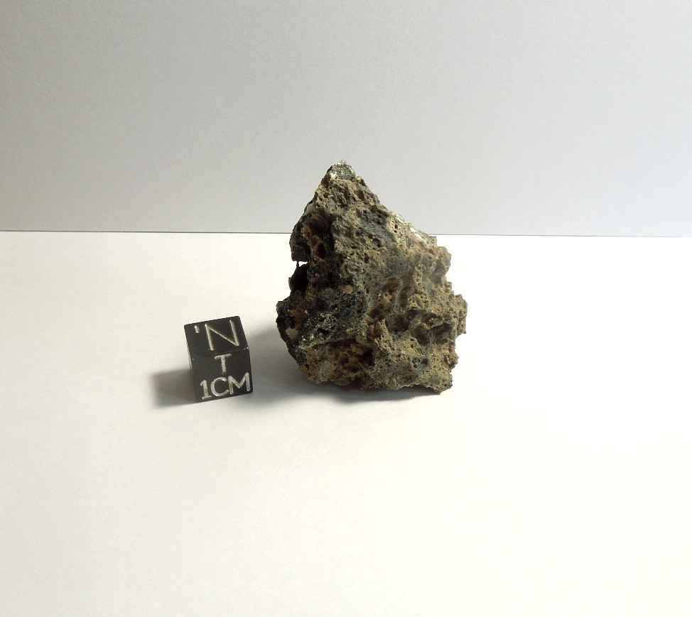 Pica Glass sample shown next to a black centimeter scale cube. The specimen is a swirled mix of grey, green, black, and white colors. The broken surface shows a highly-vesiculated interior that contains an exotic mixture of rare minerals.