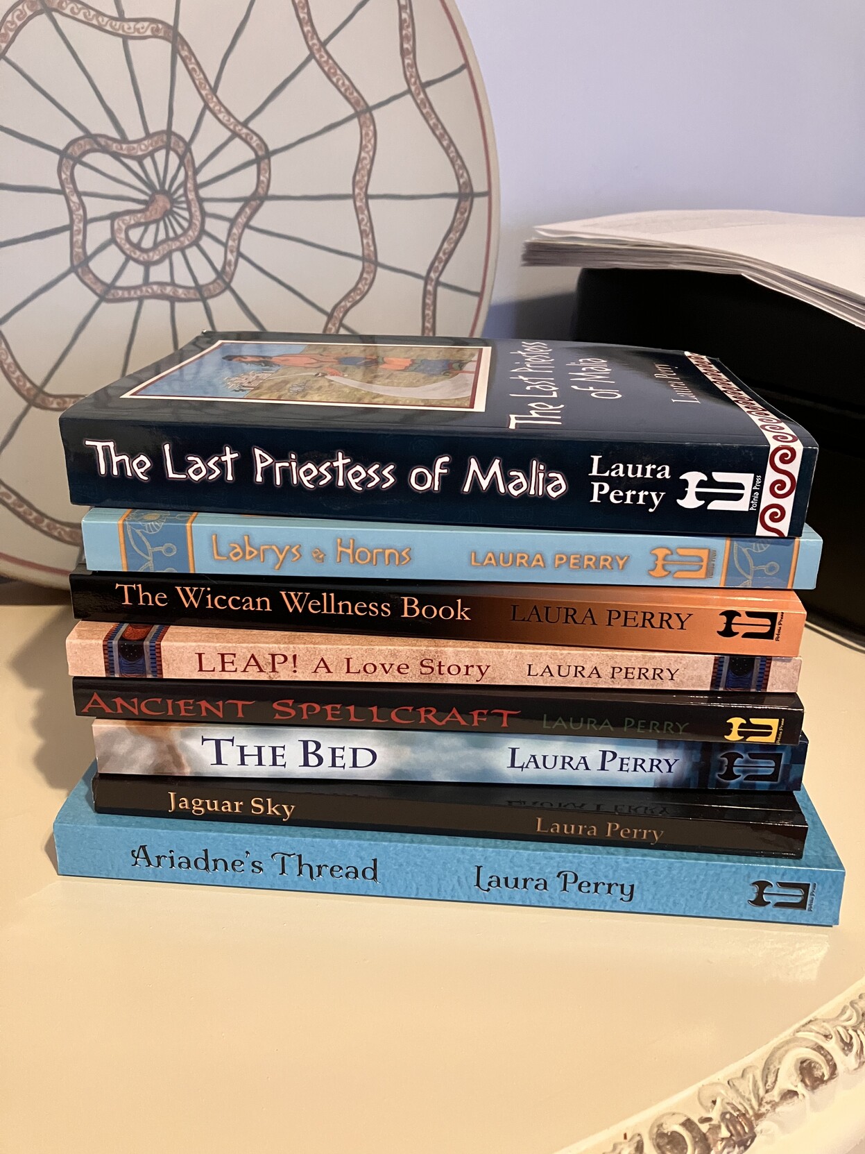 A stack of the books I've written, in paperback editions, lying on their sides with the spines facing the camera. From top to bottom the titles read: The Last Priestess of Malia, Labrys & Horns, The Wiccan Wellness Book, Leap! A Love Story, Ancient Spellcraft, The Bed, Jaguar Sky, Ariadne's Thread.