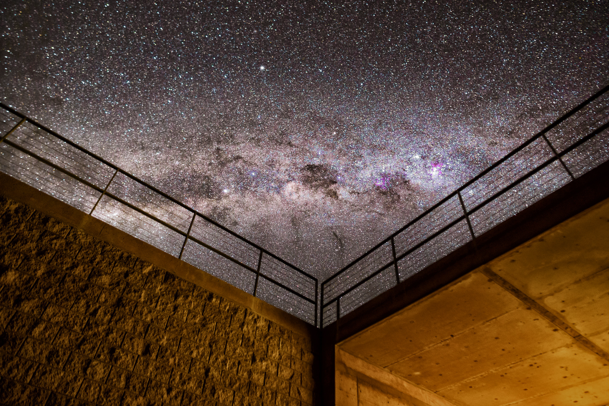 A photograph of the night sky, densely packed with thousands of stars. An even denser and brighter band of stars arches horizontally across the image, with darker patches within it. In the foreground, at the bottom of the image, two dimly-lit walls meet in a corner; the walls are topped with handrails.