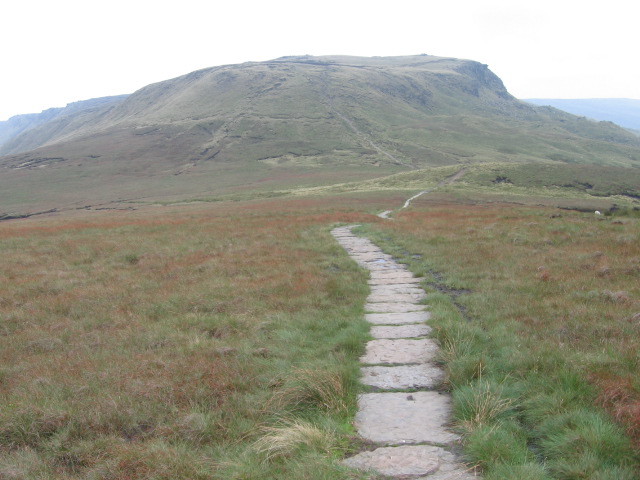 The mountain of Kinder Scout - a large, flat-topped protrusion - seen from the Pennine Way. The photo shows bleak moorland on a dark, overcast day. A paved path runs in the direction of the mountain.