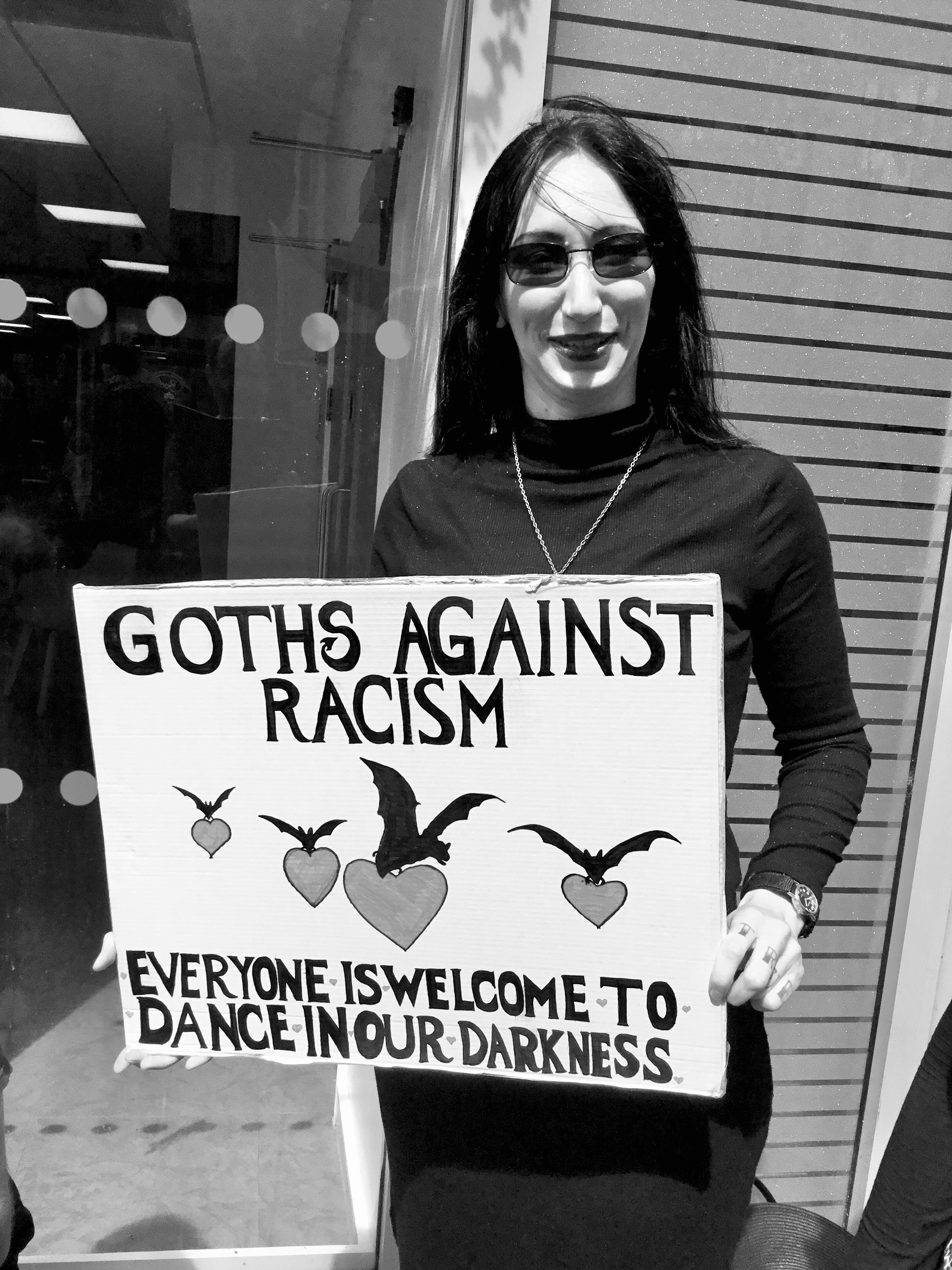 A white person in Gothic attire holds a placard that reads “GOTHS AGAINST RACISM - four bats flying in the middle carrying hearts - EVERYONE IS WELCOME TO DANCE IN OUR DARKNESS” Black and white image.