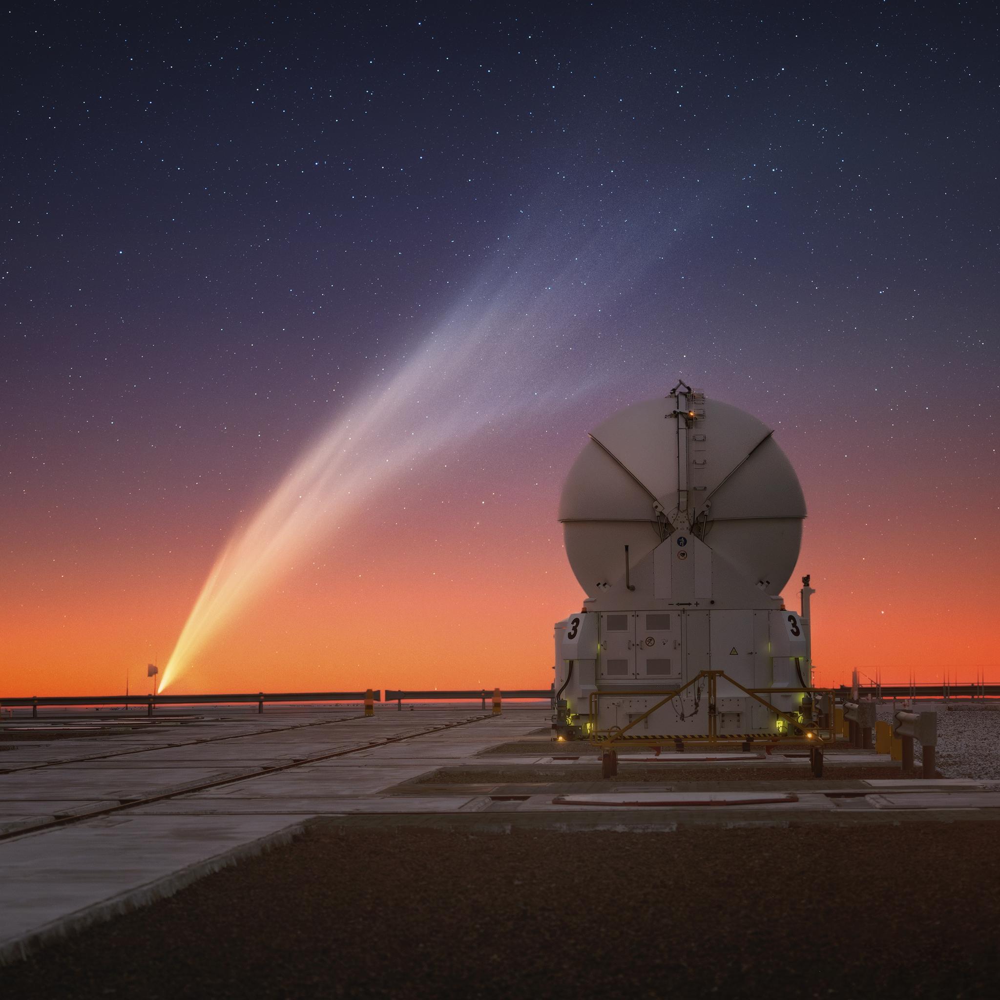 A sunset photograph at an observatory. The colour of the sky changes from blue, almost purple, to golden at the horizon. There’s a white closed telescope dome with a spherical top. To its left, the comet stretches diagonally across almost the entire image, as if it had been painted on the sky with a gigantic brush. The tail of the comet is white and full of intricate filaments.