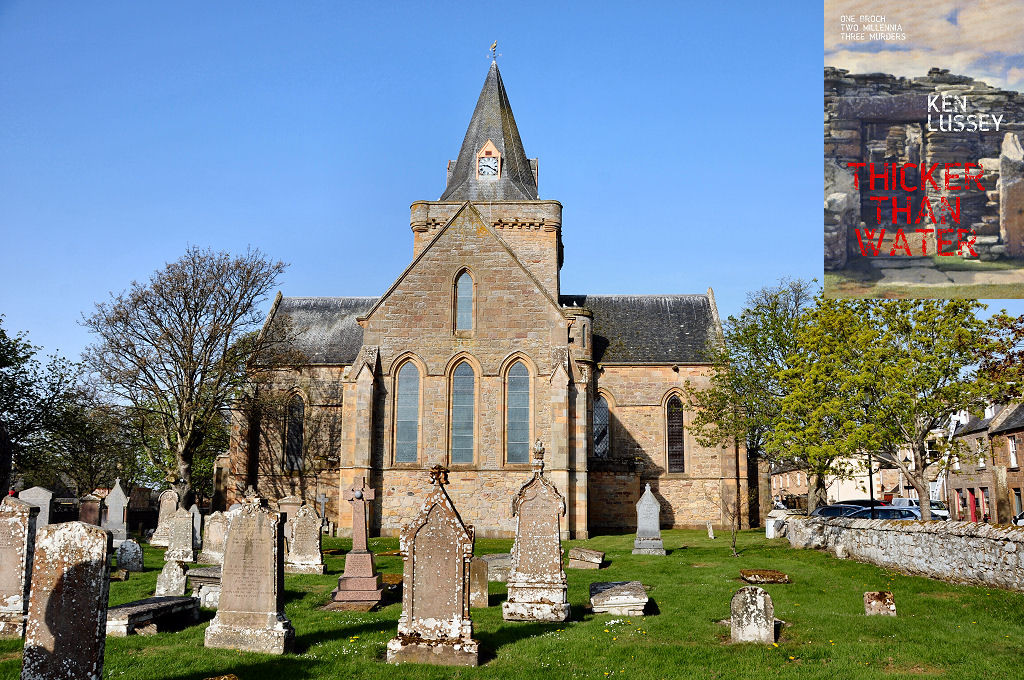 The image shows a view across a sunlit graveyard to a small honey-coloured church. We are seeing it end on, with the transepts projecting from both sides. The church is topped off with a tower and spire and there’s a low stone wall on the right with a street of stone buildings beyond it. There are trees on both sides. The sky is blue. The front cover of ‘Thicker Than Water’ is shown in the top right corner.