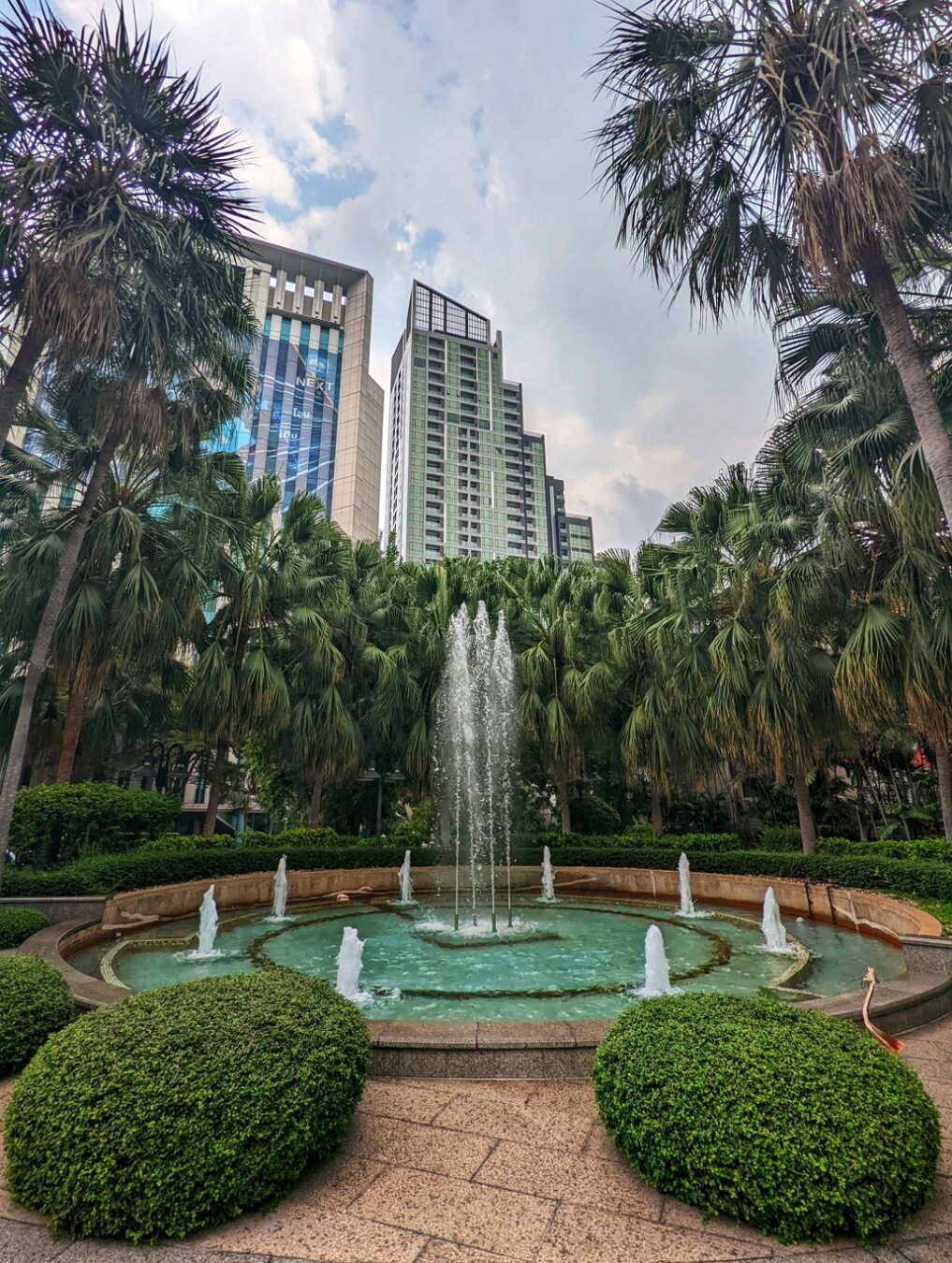 Fountain surrounded by shrubs and trees inside a garden. Tall buildings can be seen behind the palm trees. Captured with Google Pixel in Bangkok Thailand