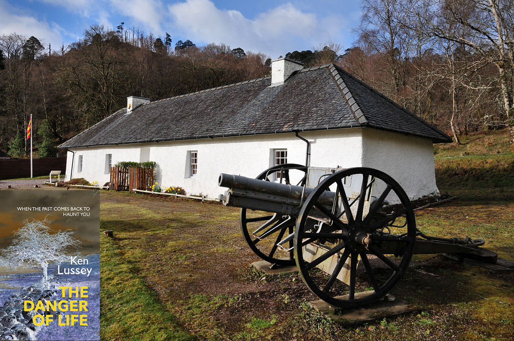 The image shows an oblique view of a long white single storey building with four windows, a central door and a pitched slate roof with two white chimney stacks. Its right end is nearest to the viewer. At the far end is a flagpole with a red and yellow striped flag while this side of the building is an old artillery piece. There are wooded hills in the background. The front cover of ‘The Danger of Life’ is shown in the bottom left corner.