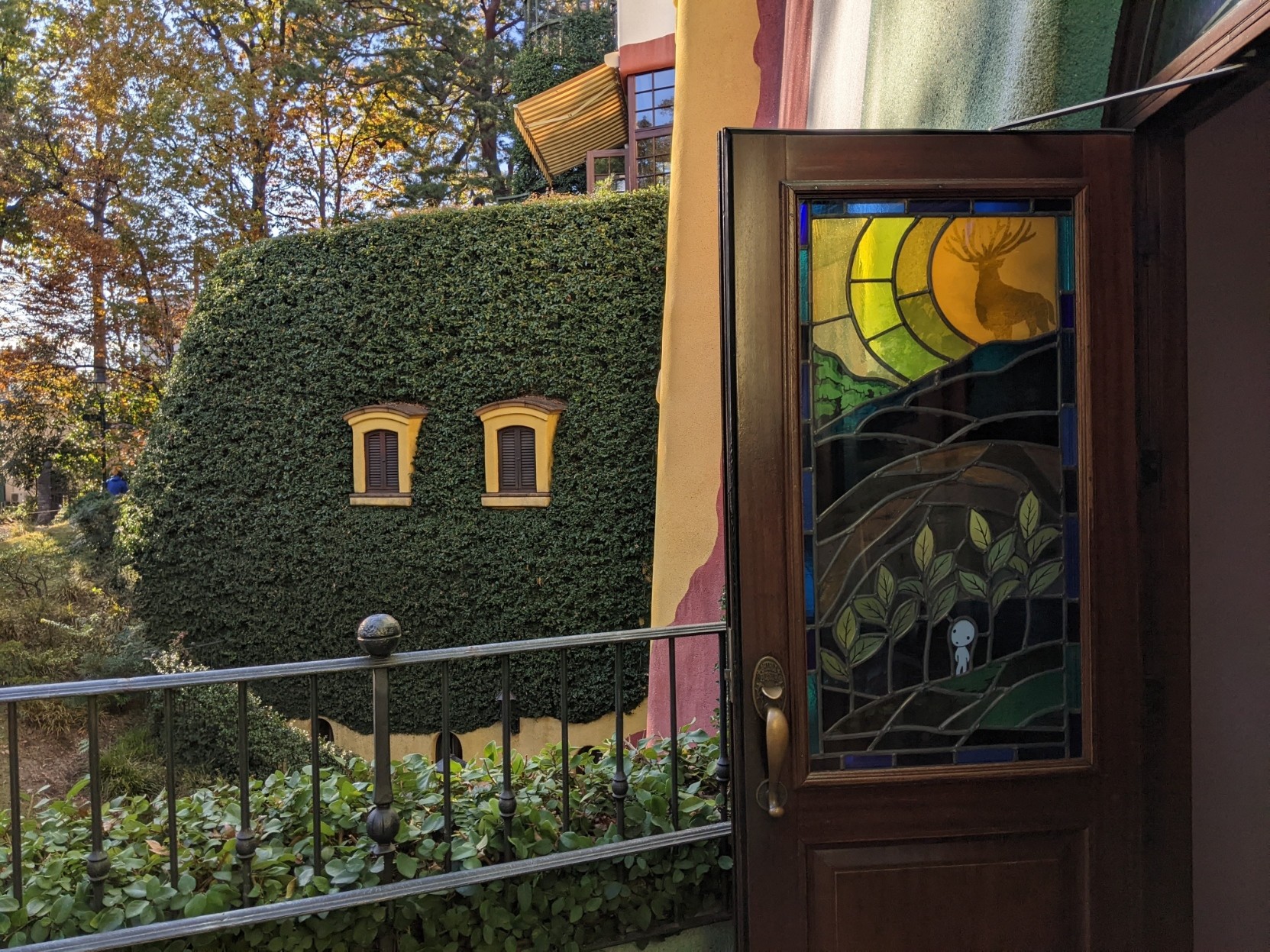 Opened Door with stained glass with Princess Mononoke art . Outside is a building wall completely covered with ivy with just 2 yellow small windows showing.