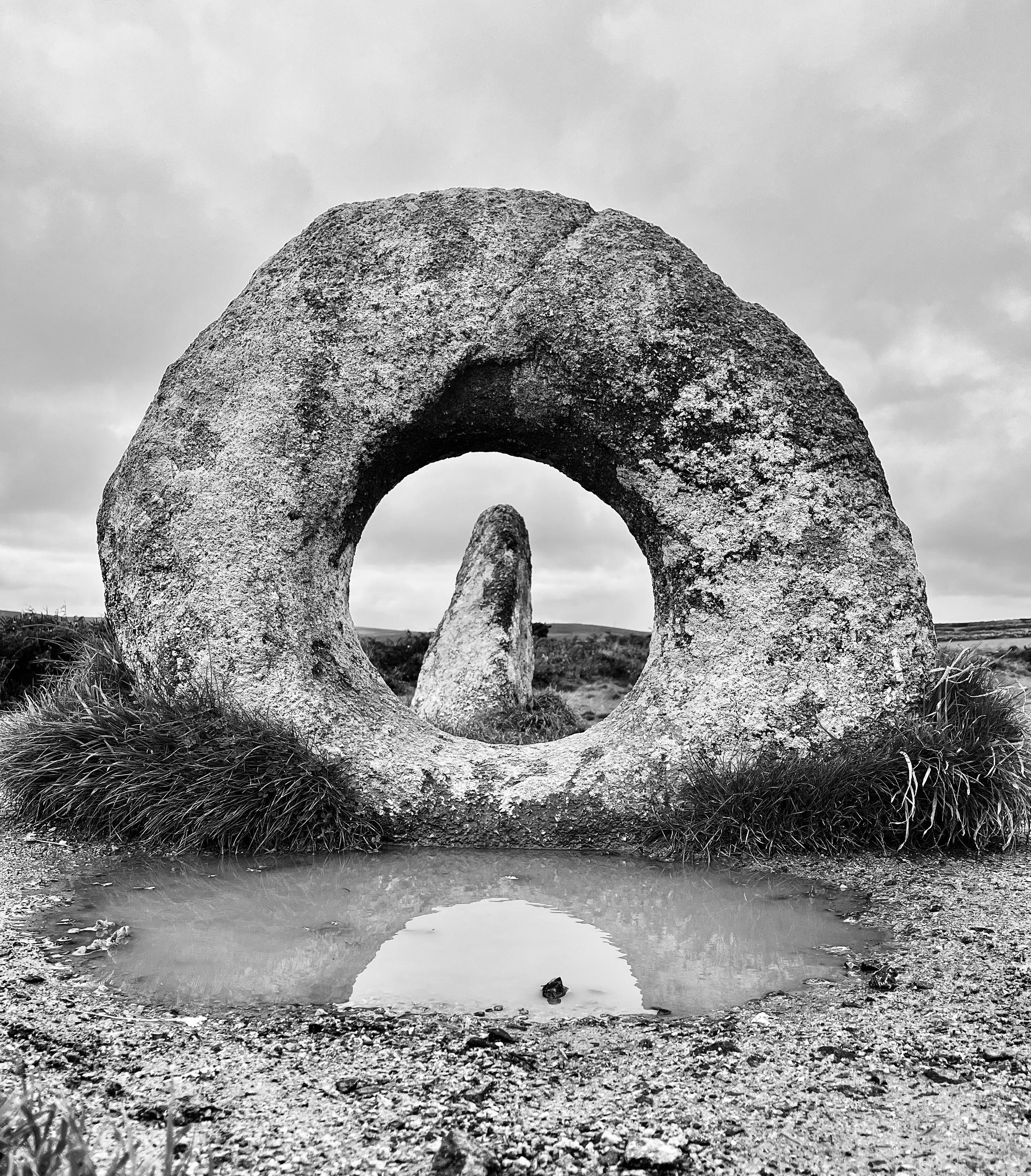 A granite round standing stone with a manmade hole in the centre. Through its aperture is framed a thin standing stone leaning slightly to the right.