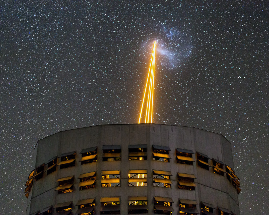 A night photograph of a cylindric telescope dome under a starry sky. Four orange laser beams leave from the telescope, converging in the direction of a dense blue cloud of stars.