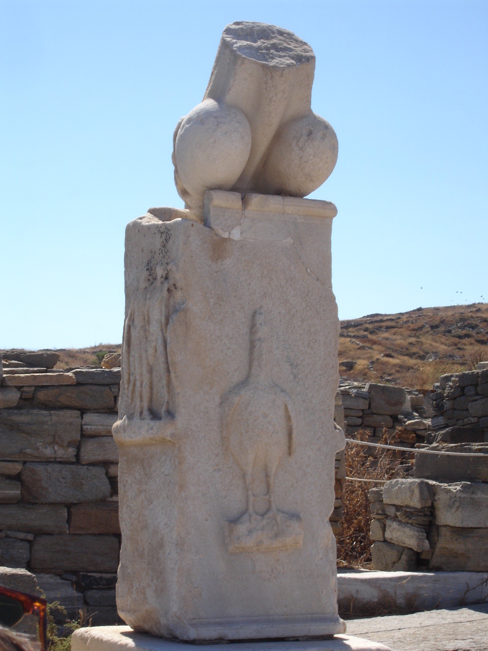 A large set of balls with the base of a shaft broken off part way through sits on a plinth with what looks like chicken facing the viewing straight on. Both phallus and plinth are marble. Photo by PhattyFatt via Wikimedia Commons.