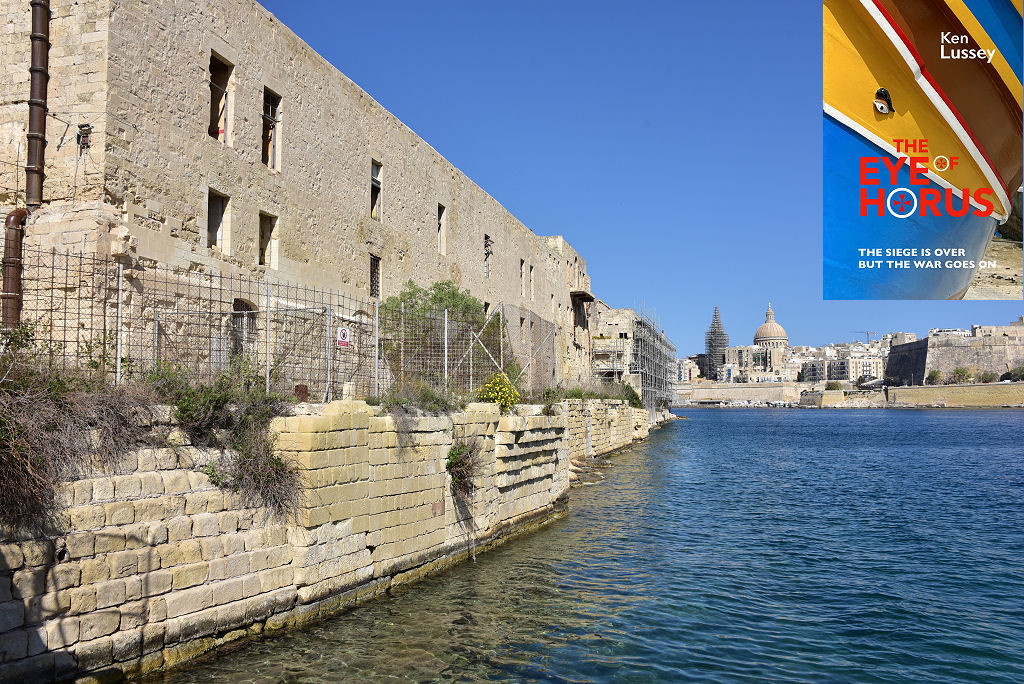 This modern image shows part of Manoel Island in Marsamxett Harbour, Malta. A light stone quayside reaches into the frame from the left, extending to the centre of the image. On the quayside are long three-storey stone buildings, fenced off and partly scaffolded. The blue water of the harbour is in the foreground and on the right and Valletta can be seen stretching across the centre of the frame in the distance. The sky is blue. The front cover of ‘The Eye of Horus’ is shown in the top right corner.