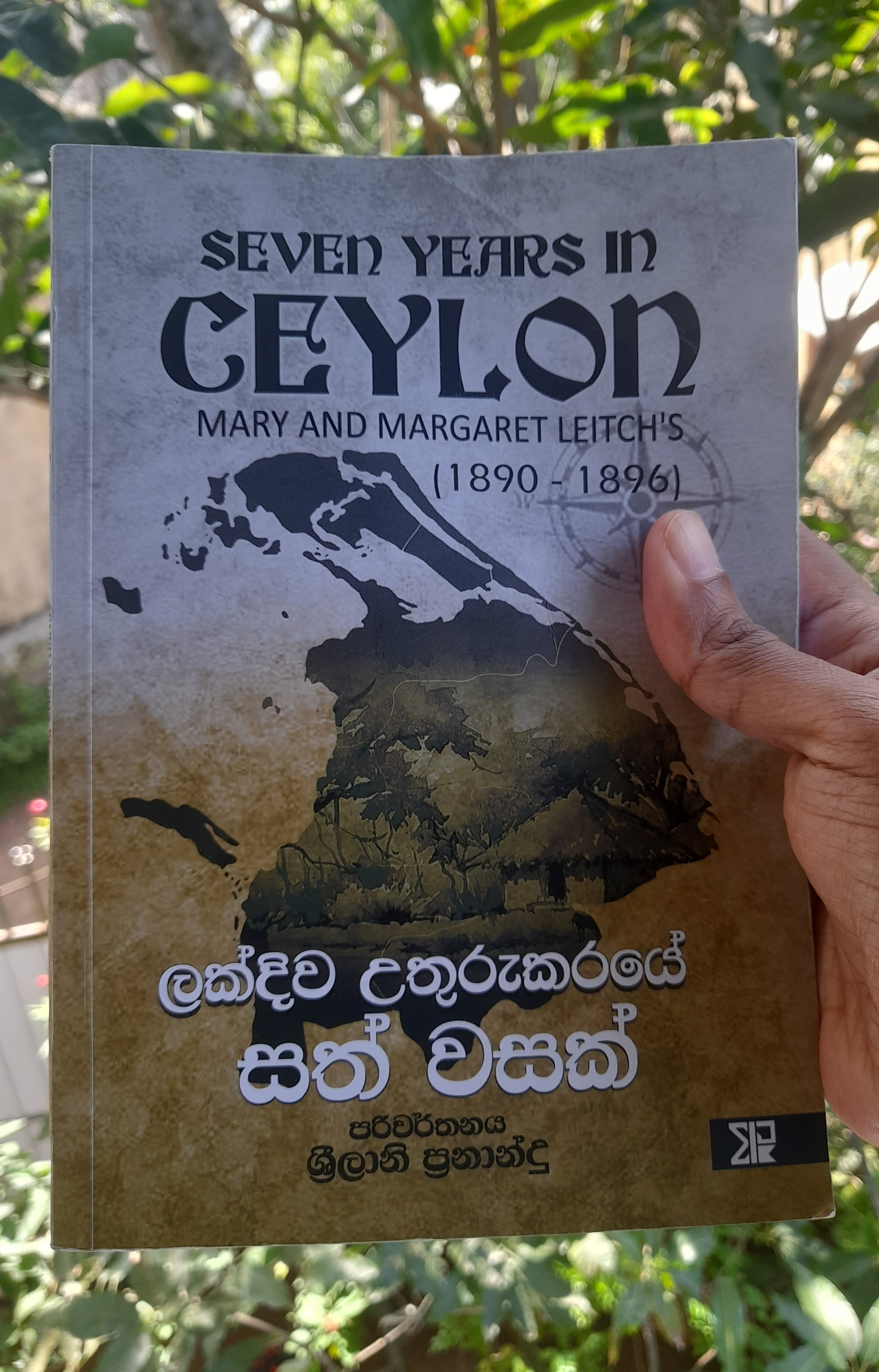 A book held in hand. The book cover of the book says "Seven Years in Ceylon" by Mary and Margaret Leitch. In the background behind the book and the hand, there is greenery lit by bright sunlight. 