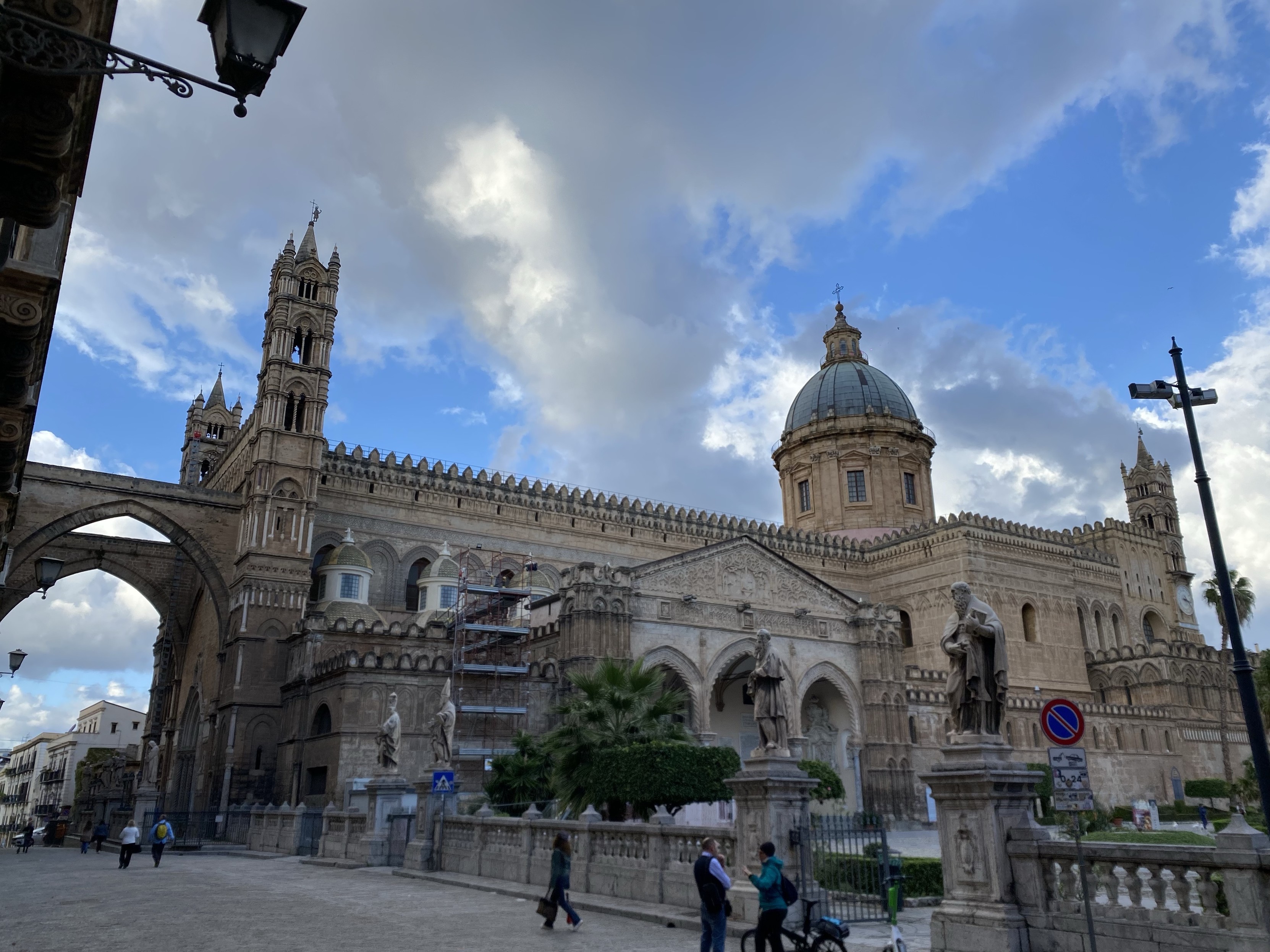 Cathedral with two bell towers and a dome against a cloudy sky, archway on the left, statues, and pedestrians walking by.