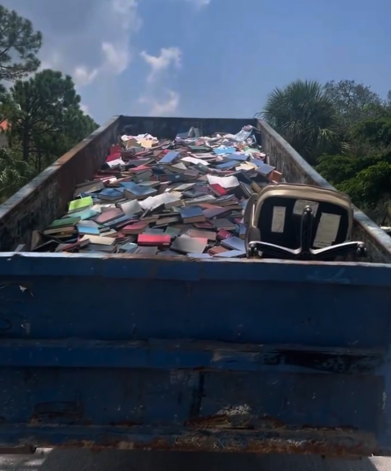 A photo of a big blue trash dumpster filled to the very top with BOOKS! :(  In one corner of the dumpster is an office chair laying  on its side. 