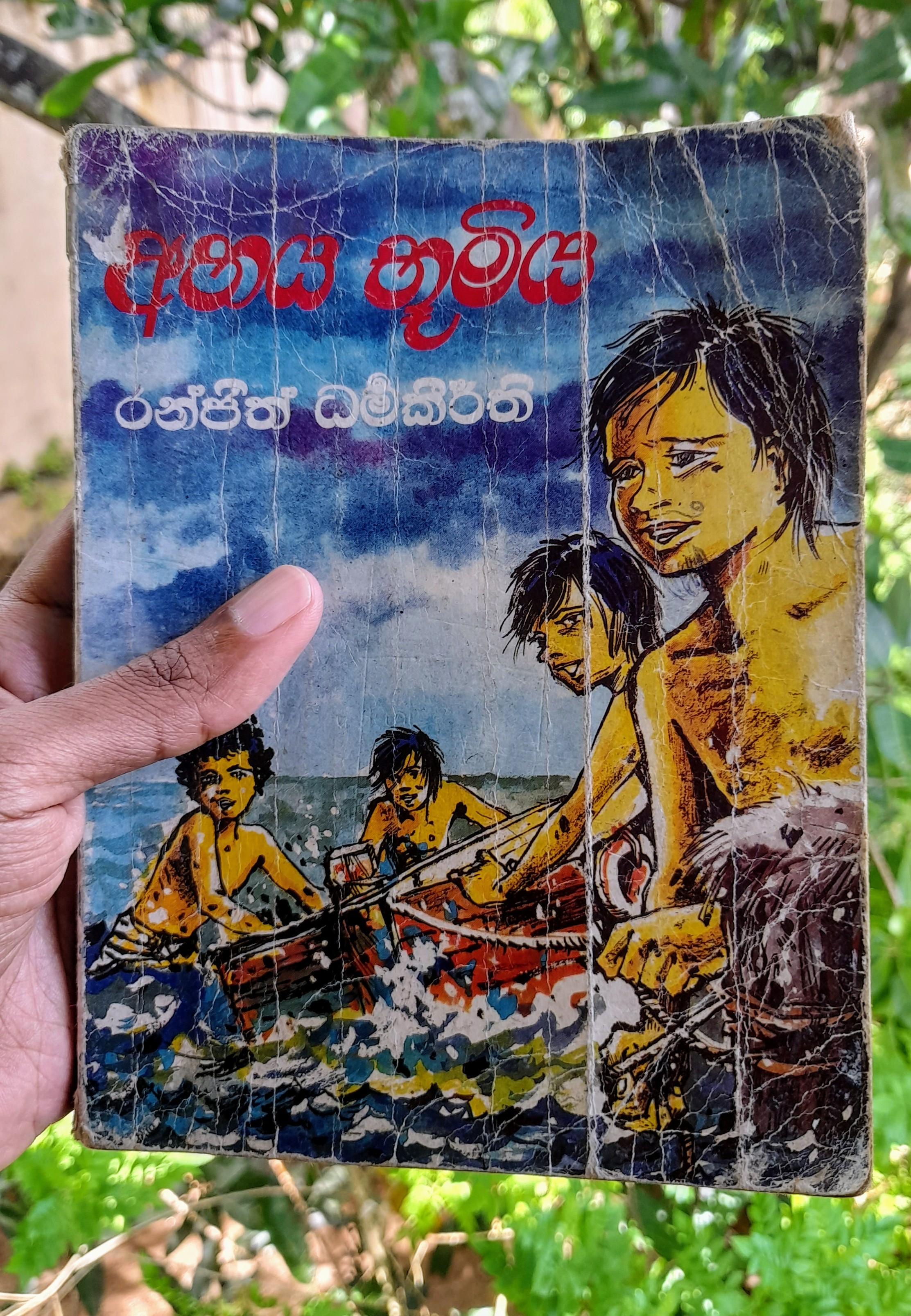 An old book held by a hand. The book cover is worn out and looks very old. The cover depicts a a drawing of 4 young boys  on water. The book cover and author name is written in Sinhala language that reads "අභය භූමිය", meaning sanctuary. The author is Ranjit Dharmakeerthi. There is greenery in the background.