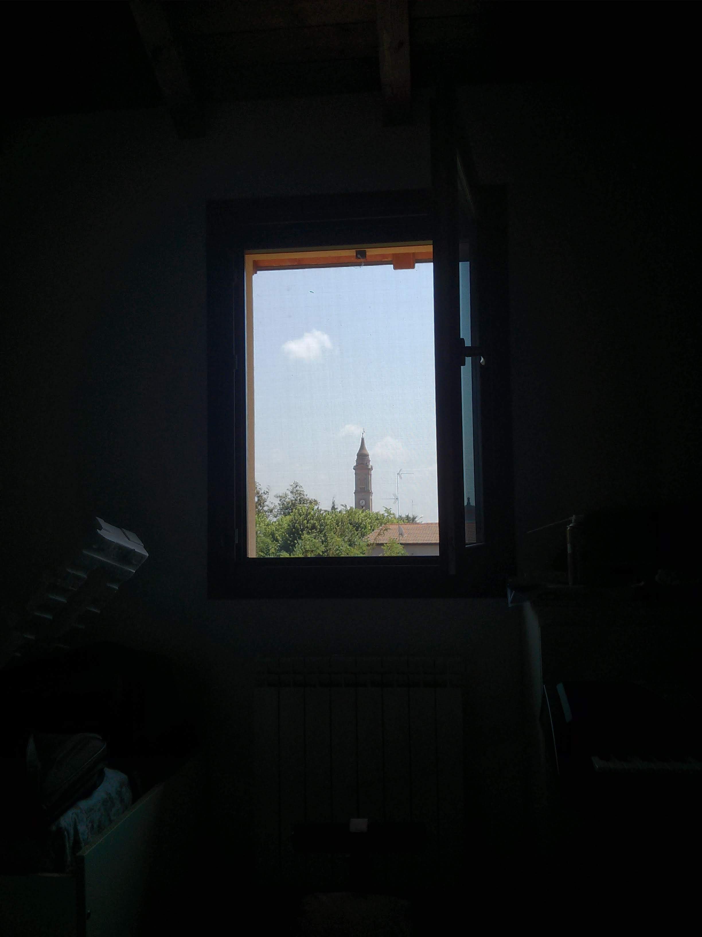 A dimly lit room with a window that brightly frames a clear blue sky. In the distance, visible through the window, is the silhouette of a towerof a church. 
The interior is too dark to make out details, but a radiator and  a piece of furniture are vaguely discernible in the shadowed foreground.