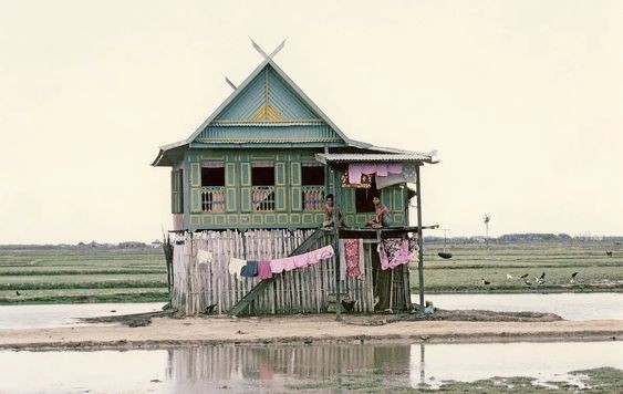 This is a photo of a typical Bugis farm house.

It shows a two-storey house surrounded by water. The house itself is on a small patch of sand.

The house has rough, undyed wood panelling on the bottom floor, with laundry strung on its outside.

The upper floor is elaborate. Many windows are visible, with colourful shutters. The upper floor is painted in a spring-green with light yellow accents. Some pink curtains are visible in each window. The house has a small balcony.

The roof is triangular, with some extensions to each side. It is panelled in light blue and with yellow accents again. The roof beams cross.

The background shows a flat, low plateau with some grass or fields. Waterbirds perch at the edge of the river. The sky is hazy with heat.

The houses are so constructed due to the Bugis belief that there are gods above and below, & that they both settled the formerly empty middle. So each floor represents a divine sphere.