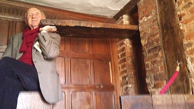 A man in late middle age holding up a false beam to reveal a hiding place. The man is in the wood-panelled library of Harvington Hall.