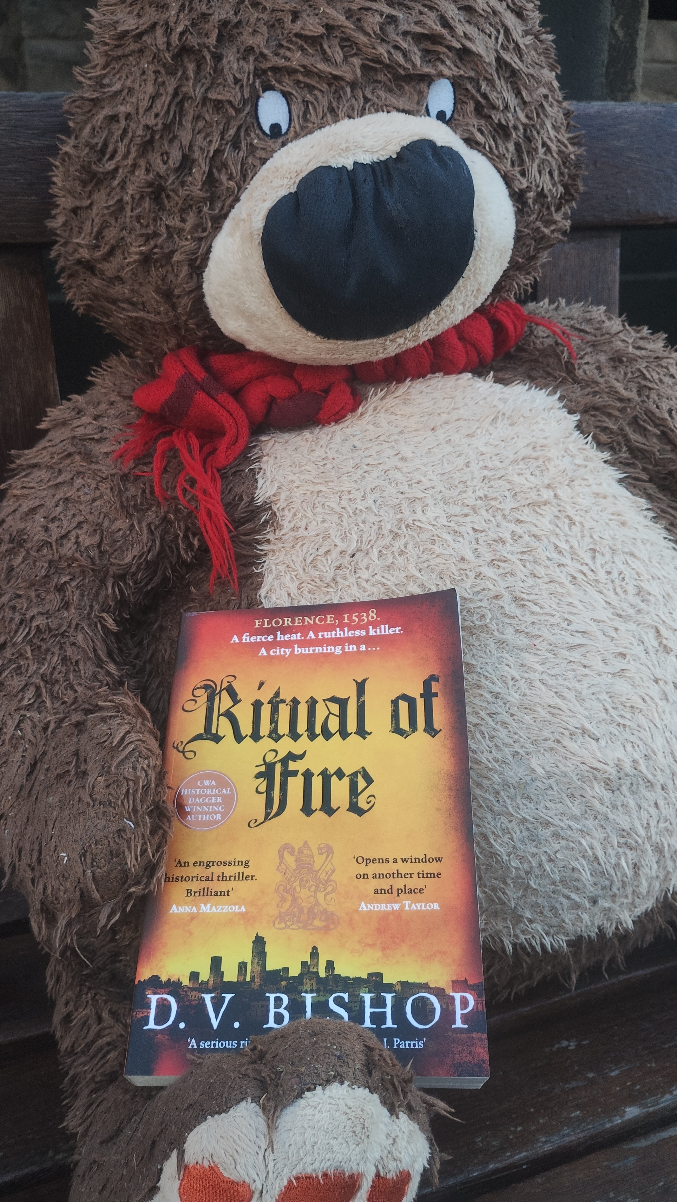 Hugless Douglass bear sitting on a park bench, holding a copy of D.V. Bishop's Ritual of Fire novel