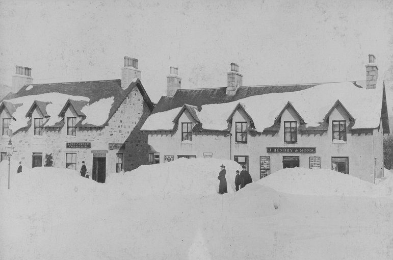 A black-and-white photo of two houses with shop fronts in Braemar. The snow is incredibly deep, and has been shoveled to allow some walkways. In front of the building on the left there is a woman and, further down, a man; and in front of the building on the right are two women and a child: the snow-drift in front of the right house is taller than the people.
Photo caption on NMS reads: 'Deep snow in Braemar, Aberdeenshire, c.1895. Reference SLA C12573. Credit: Peter M Stewart.' 