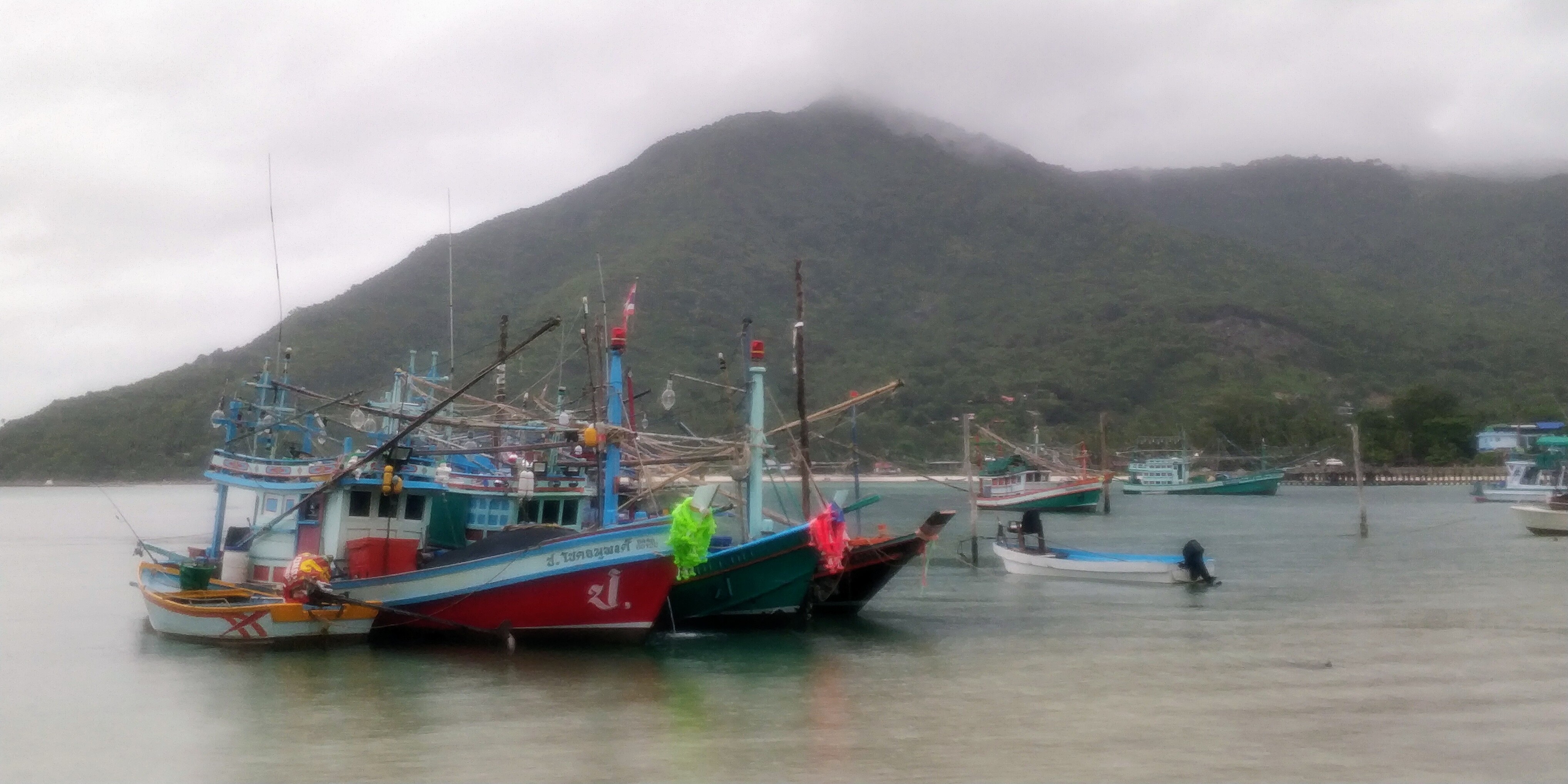  Brightly painted Thai fishing boats with a wooded "mountain" on the background, with a cloudy sky