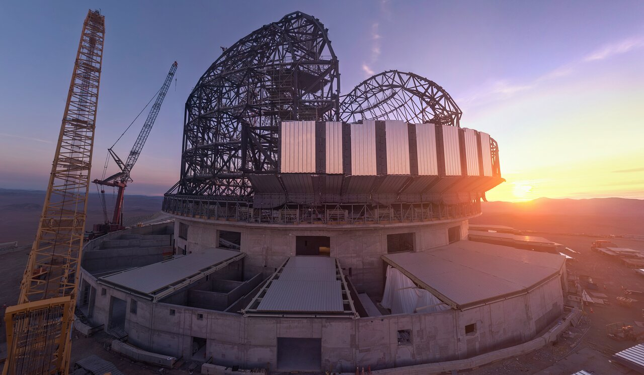 The large, steel grey skeleton of a telescope dome sits on top of a concrete foundation. At the front of the dome, light grey metal cladding covers the structure. To the right on the horizon, the yellow Sun is setting, washing the sky in shades of purple and blue. To the left, two long yellow crane arms jut into the sky.