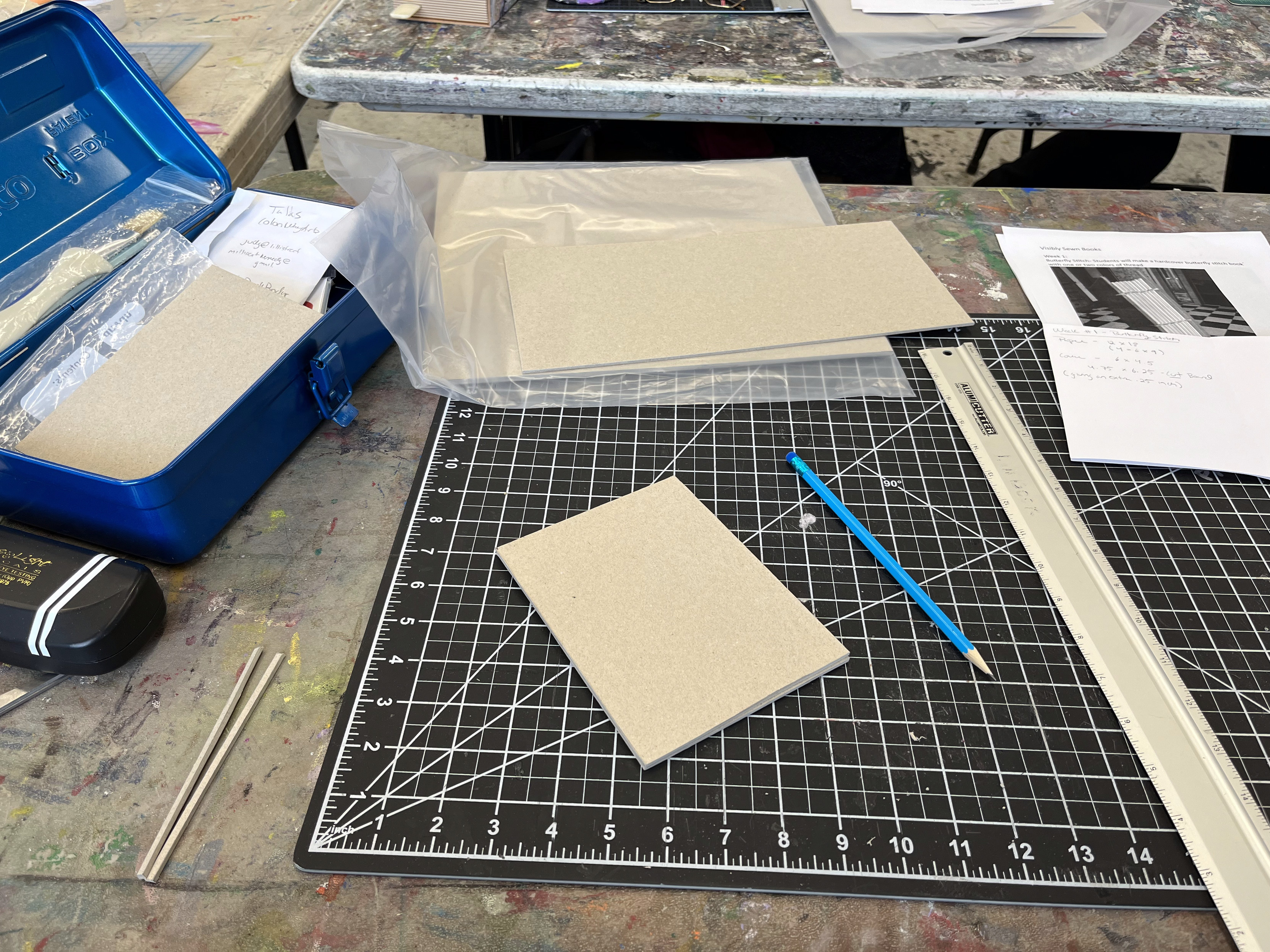 A cluttered artist's workbench with various supplies including a cutting mat, a ruler, a blue pencil, and pieces of beige board, with hints of handwritten notes and a metal box in the background.